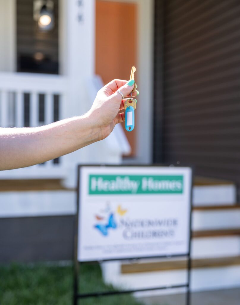woman holding a key with her home in the background; she is standing in front of a Healthy Homes yard sign